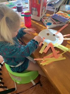 A little girl is sitting at a kitchen table making a lion mask. In front of the girl, is a paper plate with two eye holes cut out of it. The mask has yellow and orange rectangle strips glued around the edge of the mask. The little girl is still adding more strips.
