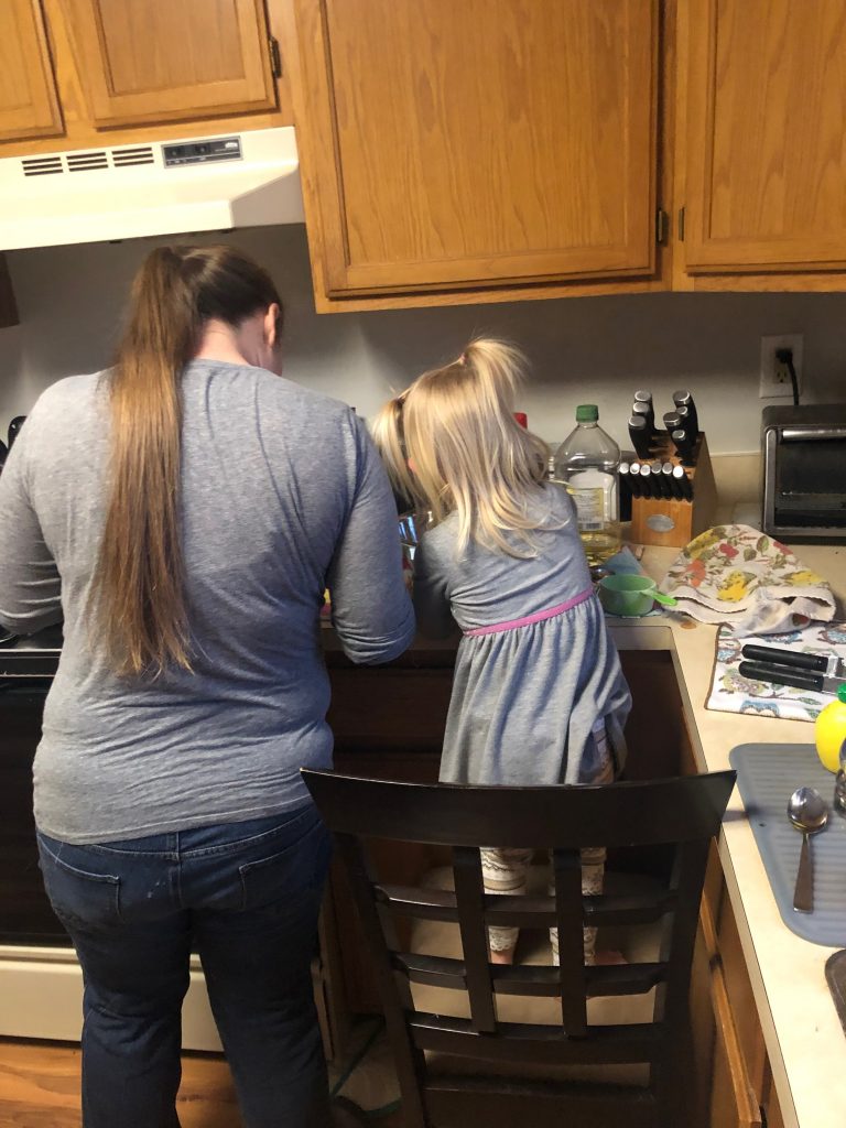 A mom is in a kitchen with a little girl, who is standing on a chair. They are standing in the corner mixing and dumping ingredients for pancakes as they practice the letter M (for measuring).