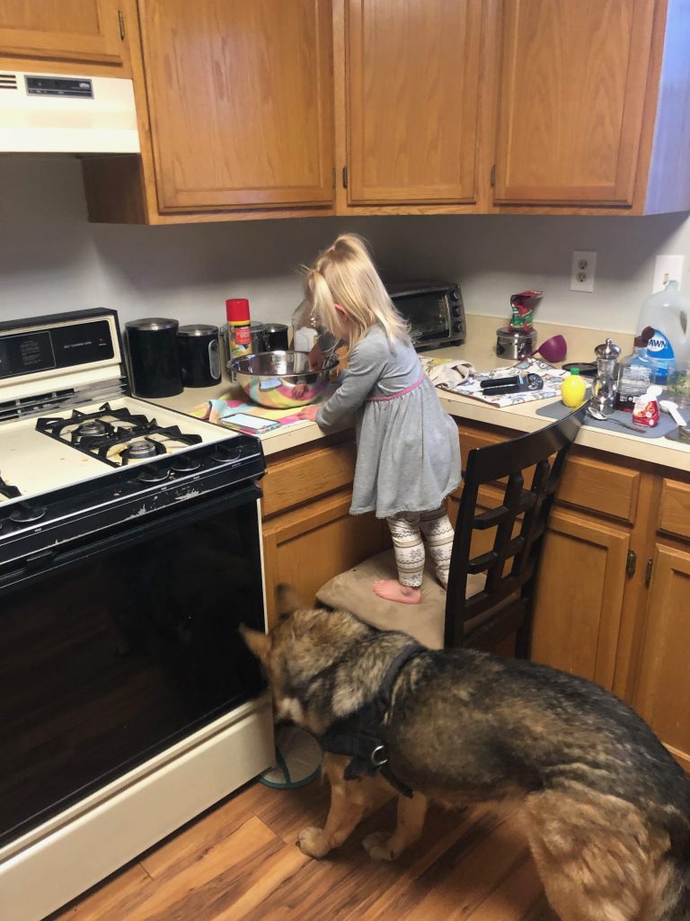 A little girl is standing on a chair in the corner of a kitchen mixing batter in a bowl as she participates in different letter M activities. This one is measuring.