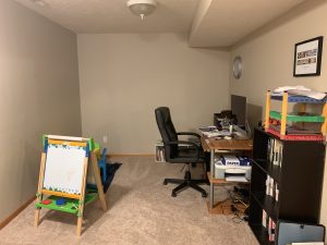 Simple homeschool organization setup in a carpeted playroom with a child-sized easel, computer desk, paper storage, cube bookshelf, books, and learning supplies.