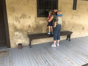 A parent and two young children explore a historical building during an outdoor learning preschool field trip. One child is carried in a front pack while the other stands on a bench, peeking through a barred window to observe inside.