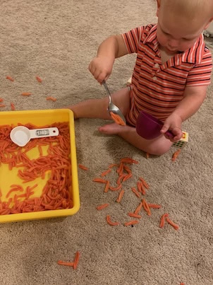 Toddler sitting on carpet scooping orange-dyed pasta from a yellow sensory bin into a purple cup using a spoon. Measuring scoops are also in the bin, showcasing toddler-friendly sensory bin products for hands-on play and fine motor skill development.