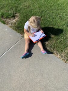 A young child sits on the sidewalk with a clipboard, drawing or writing during an outdoor learning preschool activity. She’s focused and engaged, enjoying hands-on learning in the sunshine with grass and pavement around her.