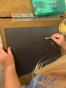 A little girl is painting with a tip on a black piece of paper.  Art is a great rainy day activity for preschoolers and toddlers.