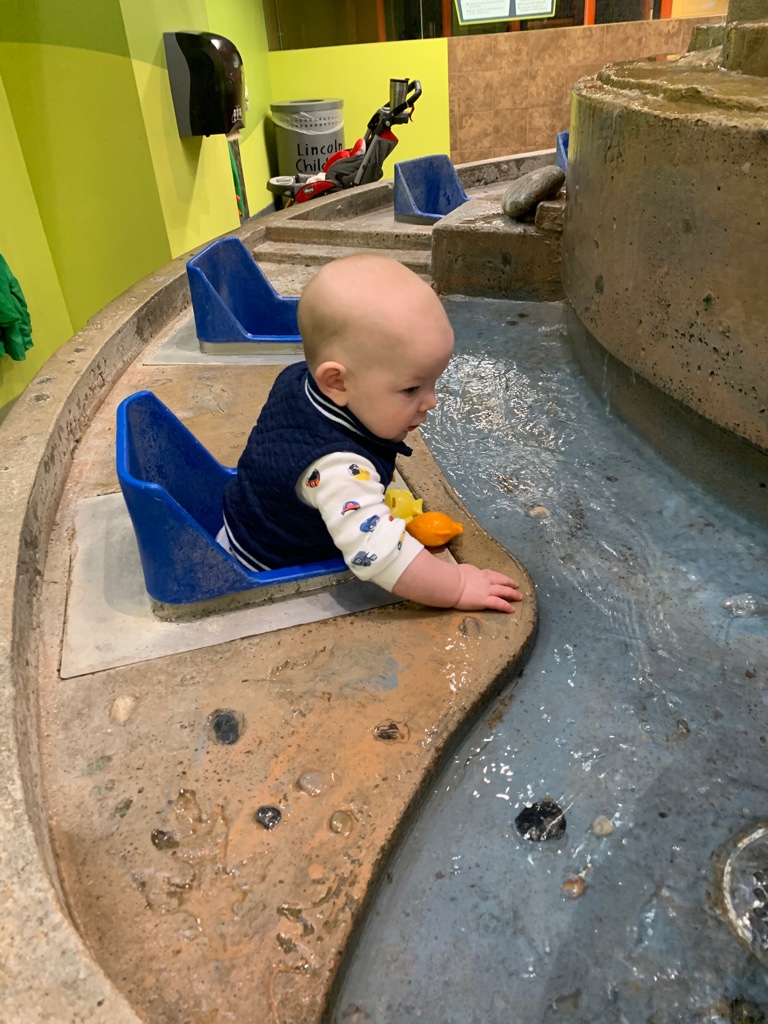 A baby boy is sitting in a chair at a water table. He is splashing in the water in front of him.
