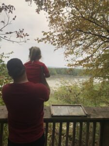A little girl is being held up on a ledge to look at water by her father on a homeschool field trip outdoors.