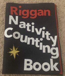 A toddler busy book on Christmas is seen laying on the ground turned to the cover. The cover reads "Riggan Nativity Counting Book."