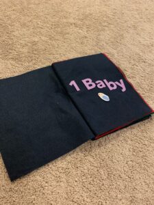 A toddler busy book about Christmas is on the floor opened to the first page. The page says "1 Baby" and has a felt baby on the page as well.