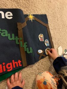 A little girl is playing with a toddler busy book about Christmas. She is placing felt people into a stable and the words, "One Beautiful Night" or seen on the other page of the book.
