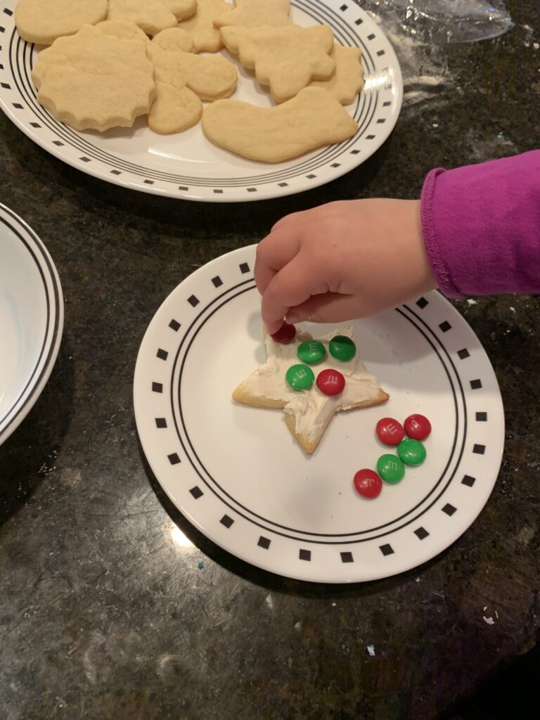 There are two plates of Christmas cookies on a counter. One plate is unfrosted and on the other plate a little girl is putting sprinkles on a frosted star cookie.