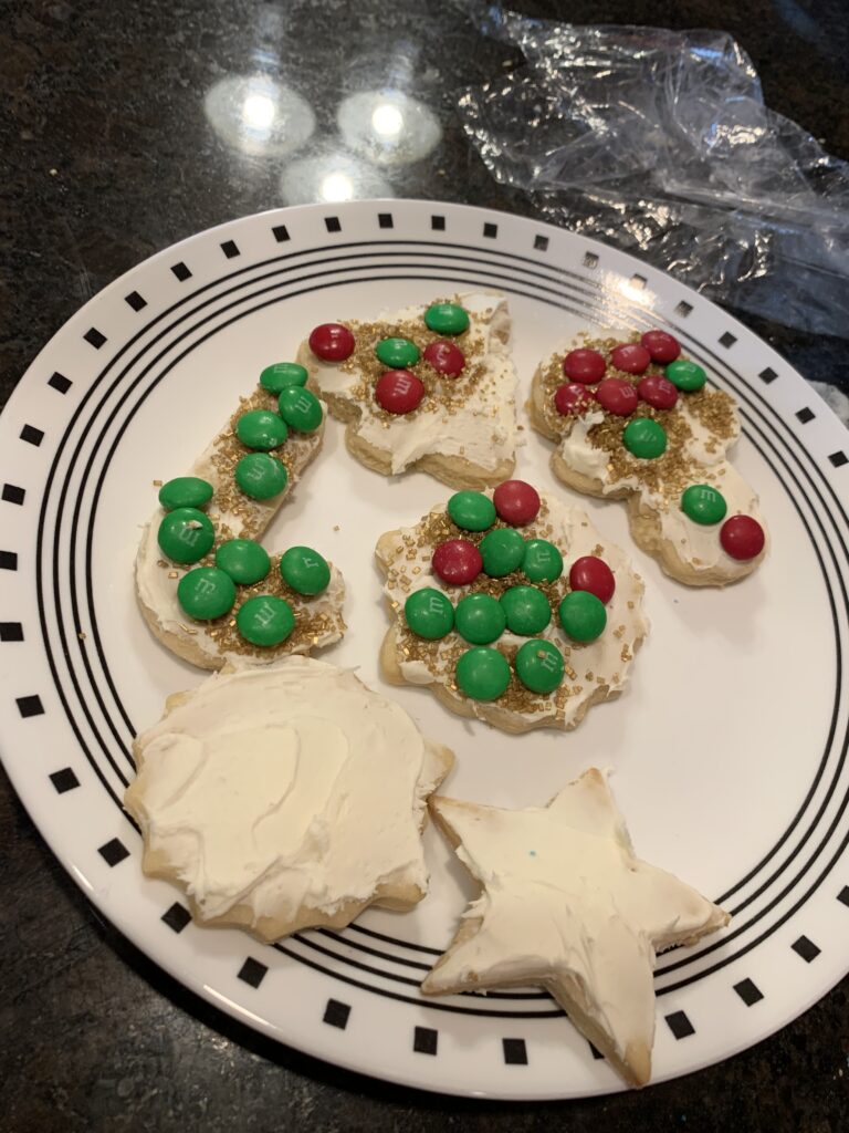 A plate of cookies is sitting on the counter. The cookies are Christmas shaped with white icing. Some of the cookies have sprinkles on them.