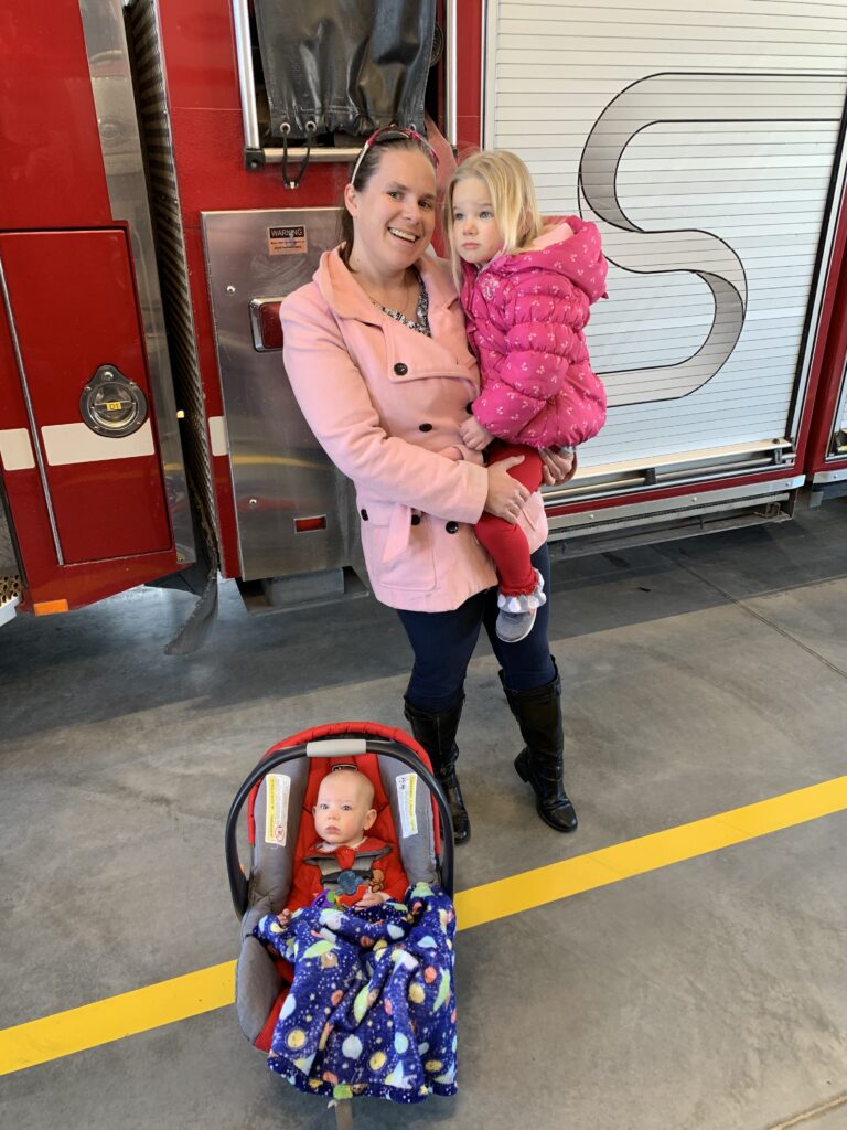 A mom is holding a little girl while standing in front of a fire truck. There is a baby boy in a car seat in front of them.