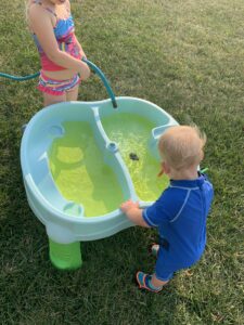 A little girl and a little boy are standing at a water table. They are filling up the water table using a hose.