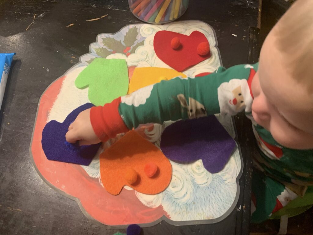 A toddler boy is sitting at a table with colored mittens in front of him.  He is placing a blue pompom on a blue mitten.