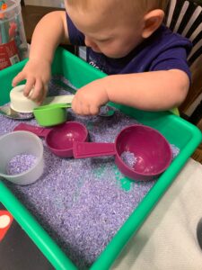 A toddler boy is sitting at a table with an open sensory bin in front of him. In the sensory bin, there is rice that has been dyed purple. There are multiple measuring cups and spoons in the sensory bin. The toddler boy is using one of the measuring cups to pick up some purple rice.