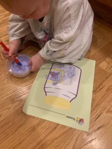 A toddler boy is sitting on the floor with a page with a jar outlined in purple in front of him. He is using a paintbrush to mix some paint, shaving cream and glue in a small cup. He has already painted some purple puffy paint on the picture.