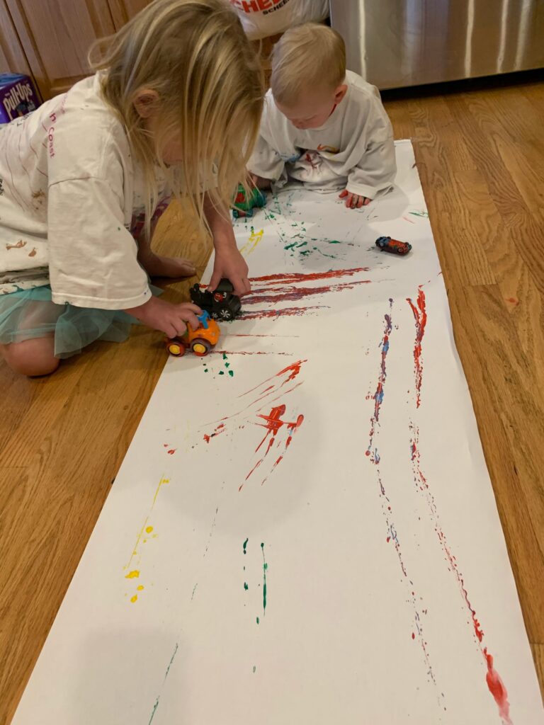 A toddler boy and a little girl are sitting on the floor of a kitchen. There is a long piece of white paper in front of them. The girl is sitting off to the side of the paper, while the toddler boy is sitting on top of the paper. Both kids have cars in their hands and they are rolling them on the paper. As they roll the cars, they are painting the paper. There are car tracks all over the paper already.