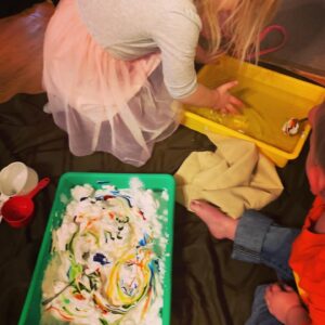 A little girl and toddler boy are sitting on a table cloth with two sensory bins on the cloth as well. The one in front of the boy has shaving cream and swirls of food coloring in it. The one in front of the girl has water in it. The girl has her hands in the water tub. The boy is dipping a spoon covered in shaving cream into the water bin. There are also an assortment of measuring cups and bowls on the table cloth near the sensory bins.