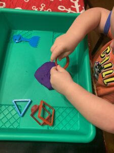 A toddler boy is sitting at a table playing with purple play dough. He is using a cookie cutter to cut a circle into the play dough. He has a few other play dough toys in his bin with the play dough.