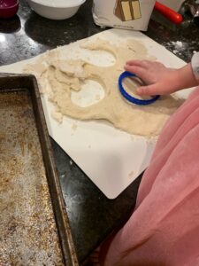 A little girl is cutting a circle out of dough. The dough is on a white cutting board where she has flattened it out. There is flour and other baking ingredients in the background and a cookie sheet in the foreground.
