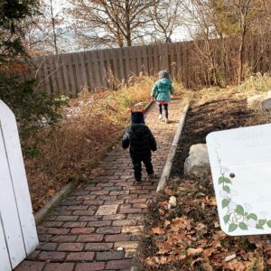 A little girl and boy are walking on a path. The path looks as though it is going through a garden that is all brown due to it being winter.
