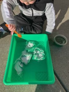 A toddler dressed in a black and gray outfit is sitting outside on a concrete surface, using an orange spoon to drop green-colored vinegar onto baking soda in a bright green tray. The mixture is creating a fizzy reaction. A small container of green liquid is nearby. This is a fun example of toddler green activities that encourage hands-on learning and sensory exploration.