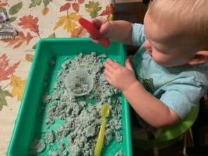 A little boy is sitting at a table, engaged in teaching colors to toddlers with a sensory bin activity. The sensory bin is filled with blue-colored flour mixed with oil, perfect for teaching colors toddlers blue in a hands-on way. The boy is using a spoon to scoop the blue flour into a measuring cup, making this activity ideal for teaching colors toddlers blue through sensory play.