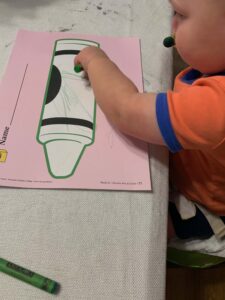 A toddler is coloring a large crayon outline on a pink worksheet using a green crayon. The child is wearing an orange and blue shirt and is focused on the activity. Another green crayon rests on the table nearby. This is a great example of toddler green activities that help develop fine motor skills and color recognition.