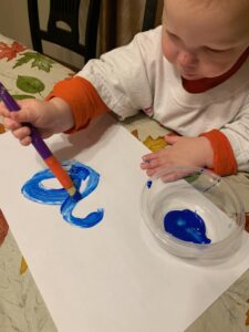 A toddler boy is sitting at a table painting, actively engaged in teaching colors to toddlers with a focus on blue. In front of him is a blank piece of paper, and a small container of blue paint sits beside it, emphasizing teaching colors toddlers blue through art. Using a jumbo paintbrush, he is creating random strokes of blue paint on the paper, making this a fun and effective activity for teaching colors toddlers blue.
