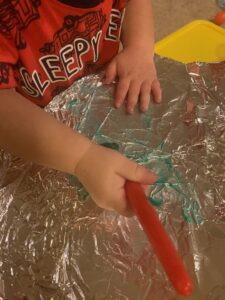A toddler boy is sitting at a table painting with a paintbrush. His activity is focusing on teaching colors toddlers blue. Instead of paper, the boy is painting on foil, making it a unique activity for teaching colors toddlers blue. There is a bit of blue paint on the foil, highlighting how teaching colors toddlers blue can be both creative and fun.