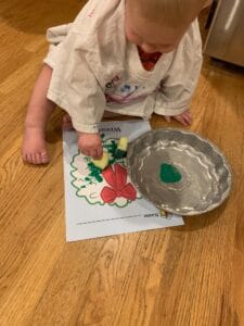 A toddler wearing a paint shirt is painting a picture of a wreath with a sponge and green paint. The green paint is in a foil pie pan next to the child. This is one of many great green activities for a toddler.