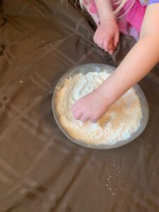 There is a little girl sitting on a table clothe laid out on the floor.  She has her hand in a pan of flour making moon craters with rocks.