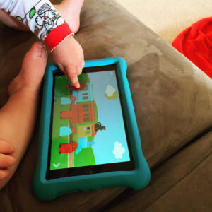 A toddler boy is sitting on a couch with a tablet in front of him. He is playing a Lego game on the tablet and moving a Lego with his finger.