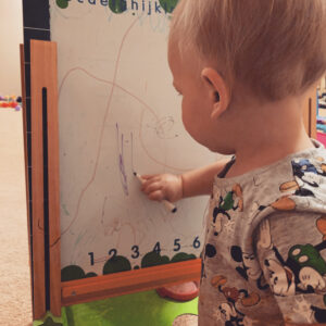 A toddler boy is standing in front of a child's art easel. He is using a dry erase marker to color on the dry erase board.