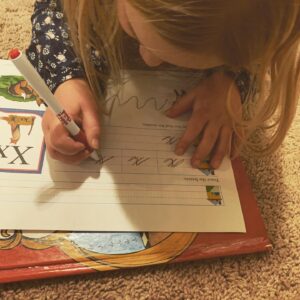 A little girl is laying on the floor writing on a worksheet on a book. On her worksheet, she is tracing cursive x letters with a pen.