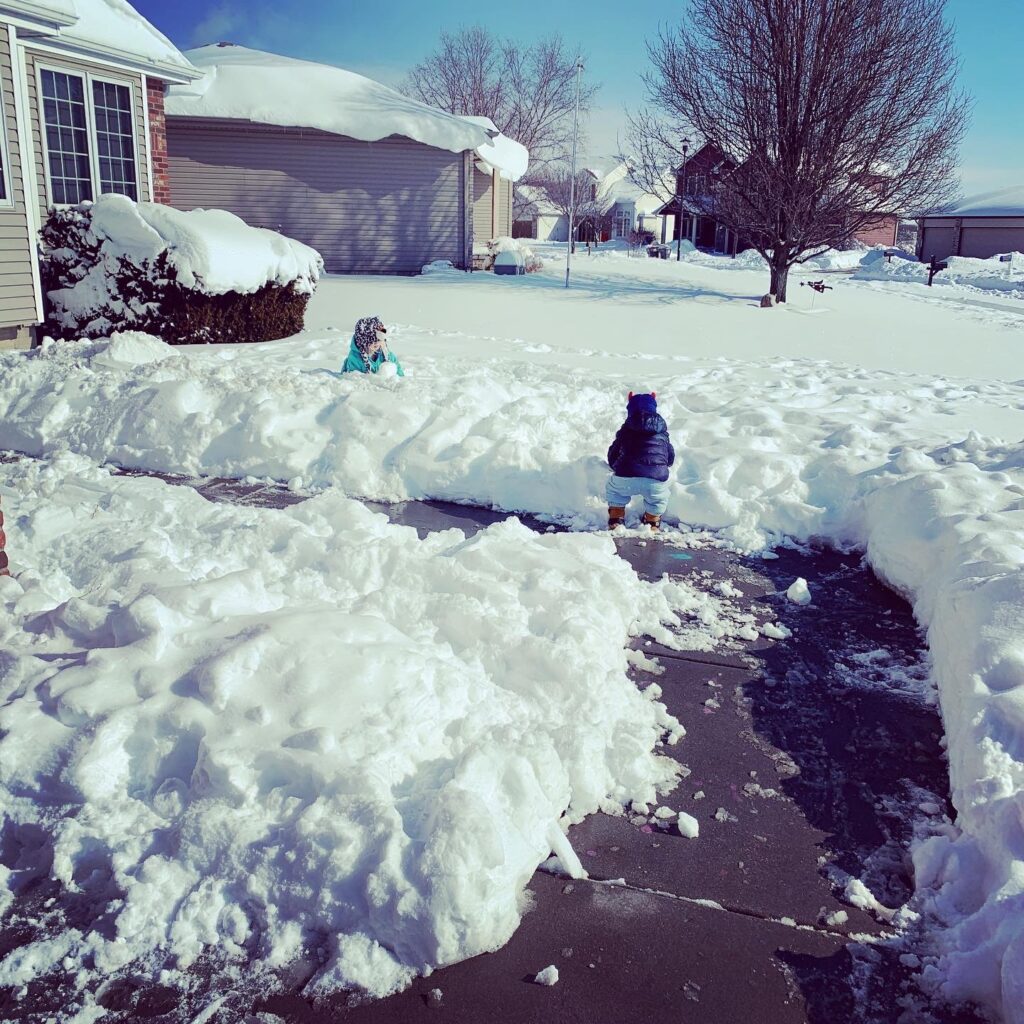 A little girl is sitting in a yard full of snow making snowballs. There is a toddler boy on the sidewalk playing int he edge of the snow.