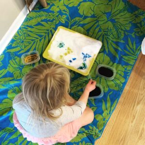 A little girl is sitting on a flowery towel with a bin of snow in front of her. Beside her are three small containers of colored water (two on one side and one on the other side). The little girl is using a pipette to drop colored water onto the snow.