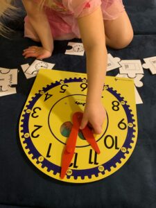 A little girl is sitting on a couch with puzzle pieces all around her. She is moving the hands on a Judy clock to make 11 o'clock.