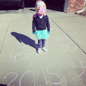 A preschool-aged child participates in an outdoor learning activity, standing on a sidewalk chalk number line counting by fives. She's wearing a helmet, jacket, gloves, and tutu, engaging in playful math exploration during outdoor learning preschool time.