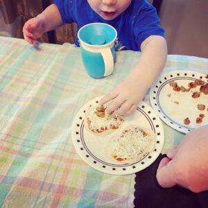 A toddler boy is sitting at a dinning room table putting toppings on mini pizzas.  He has his water cup in front of him and he is placing sausage on the pizzas on a plate on the table.