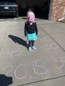 Preschooler playing an outdoor skip counting game with chalk numbers on the driveway, combining movement and play-based learning for early math development.