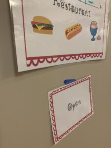 The dramatic play restaurant sign is hanging on the wall.  You can see a young child has written on it.  The open sign hangs underneath the restaurant sign.