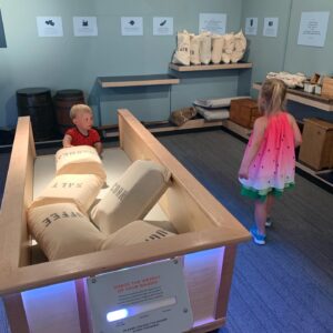 A little boy and girl are playing at a museum. The girl is walking around looking for something to load into a pretend wagon. The boy is loading a bag of goods into the wagon. They are trying to see how much they can take on a trip like a pioneer would take.