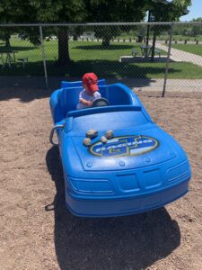 A little boy is sitting in a fake car at a playground. The car is blue and has rocks on the "hood." He is rocking the car forward and backwards while turning the fake steering wheel.