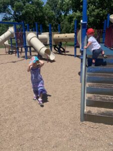 A boy is climbing stairs on a playground while a little girl walks towards the stairs fixing her baseball cap.