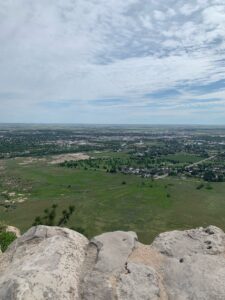 This is the view from on top of Scotts Bluff National Monument. There are rocks on the bottom of the picture that show the edge of the cliff. Out past the cliff you can see grass and a city in the distance.