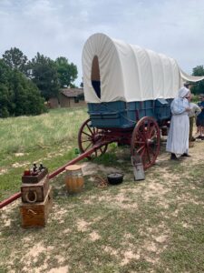 A wagon made to look like a wagon on the Oregon trail is the main part of the image. There are boxes with items from that time period are around the wagon. There is also a reenactor dressed like a pioneer woman in front of the wagon.