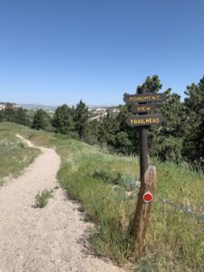 There is a trail marker on the right side of the image that says, "Monument View Trailhead." There is a gravel trail leading off to the distance next to the sign with grass and tress surrounding it.