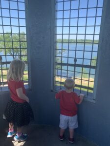 A little boy and a little girl are standing inside a lighthouse looking out of the top. There are windows in front of them with bars on the windows. Outside the windows you can see the water of the lake and trees and grass around the lake.