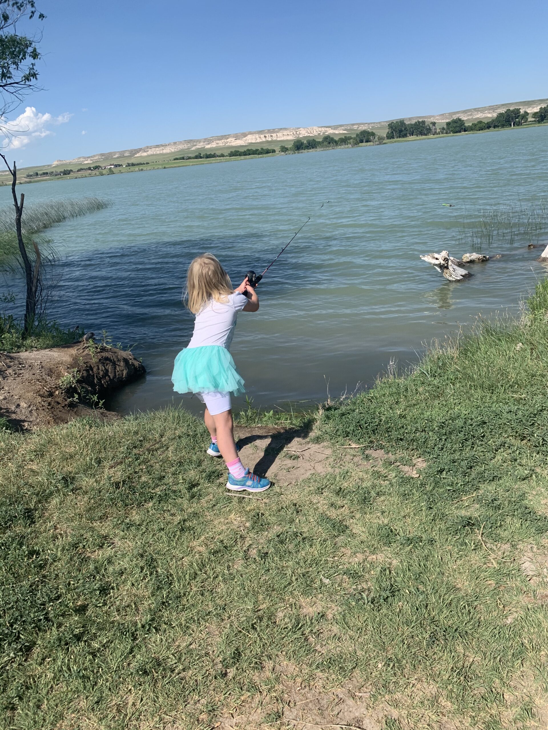 A little girl is casting out her fishing rod into a lake. She is standing on the shore right at the edge of the lake.